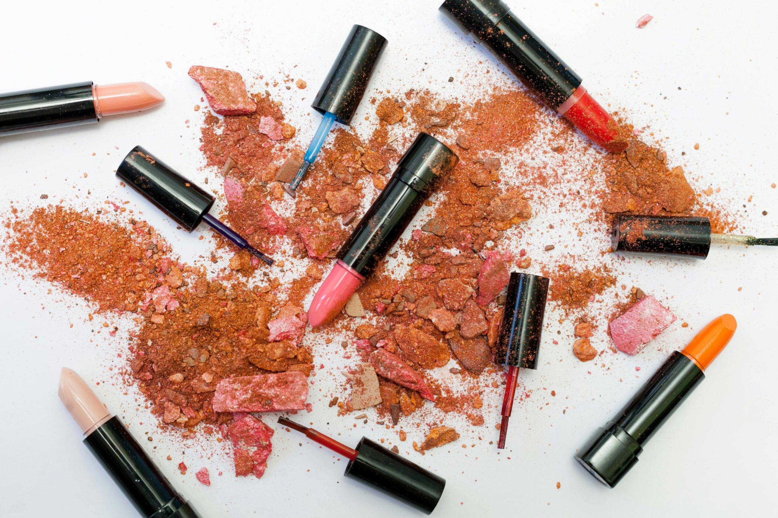 Colorful scattered makeup products including lipsticks and crushed powders on a white background.