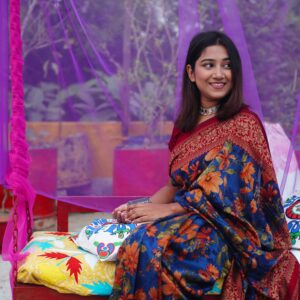 Young woman wearing a colorful floral saree, sitting outdoors with a joyful smile.